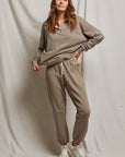 A person wearing the French Terry Easy Sweatpant from Perfectwhitetee in a buttery soft taupe shade pairs it with white sneakers, standing against a light gray backdrop. They have long hair and are casually posed with one hand holding the hem of their french terry cotton sweatshirt. The outfit embodies a relaxed, comfortable style.