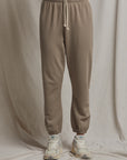 A person wearing the French Terry Easy Sweatpant by Perfectwhitetee, designed in ankle-length with a drawstring and made of French terry cotton, stands against a neutral beige fabric background. They are also wearing white and beige sneakers, highlighting an elevated comfort wear sense. The image emphasizes the lower half of the body.