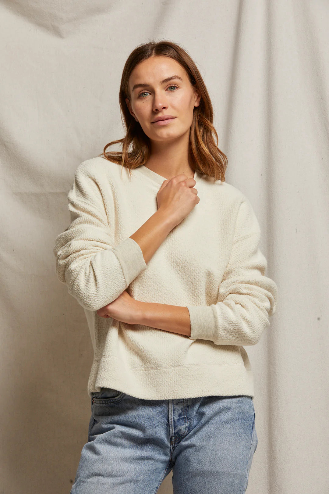 A woman with shoulder-length brown hair poses casually in a Mayer LS reverse fleece by Perfectwhitetee. She stands against a neutral fabric background, wearing blue jeans and looking towards the camera with a subtle smile, exuding an effortlessly vintage look.