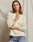 A woman with shoulder-length brown hair poses casually in a Mayer LS reverse fleece by Perfectwhitetee. She stands against a neutral fabric background, wearing blue jeans and looking towards the camera with a subtle smile, exuding an effortlessly vintage look.