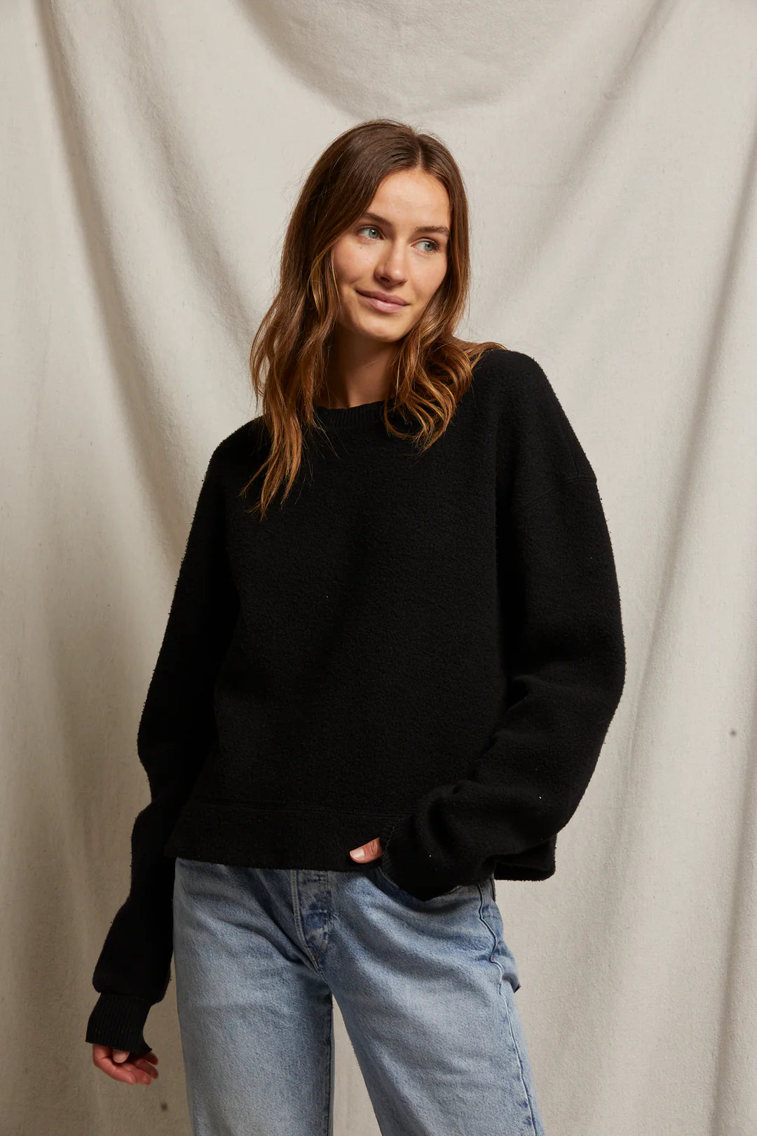 A person with long brown hair, dressed in the "Mayer LS reverse fleece" by Perfectwhitetee, paired with blue jeans, stands before a light-colored fabric backdrop, embodying a vintage look and glancing to the side with a slight smile.