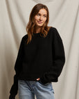 A person with long brown hair, dressed in the "Mayer LS reverse fleece" by Perfectwhitetee, paired with blue jeans, stands before a light-colored fabric backdrop, embodying a vintage look and glancing to the side with a slight smile.