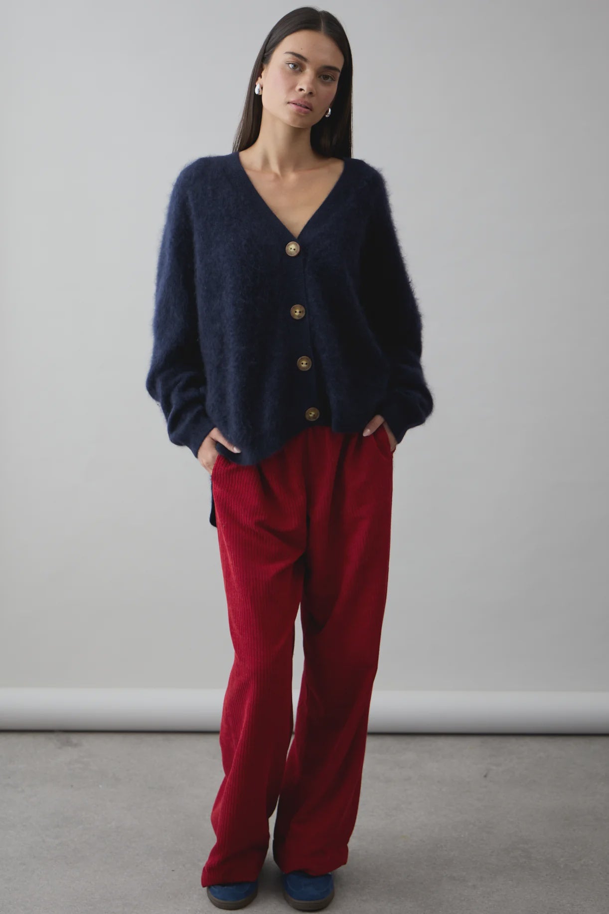 A woman stands against a plain background, hands in her pockets, calmly looking at the camera. She wears the dark blue Mae Cardi by Mia Fratino Cashmere, loose red corduroy pants, and blue shoes.