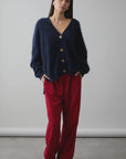 A woman stands against a plain background, hands in her pockets, calmly looking at the camera. She wears the dark blue Mae Cardi by Mia Fratino Cashmere, loose red corduroy pants, and blue shoes.
