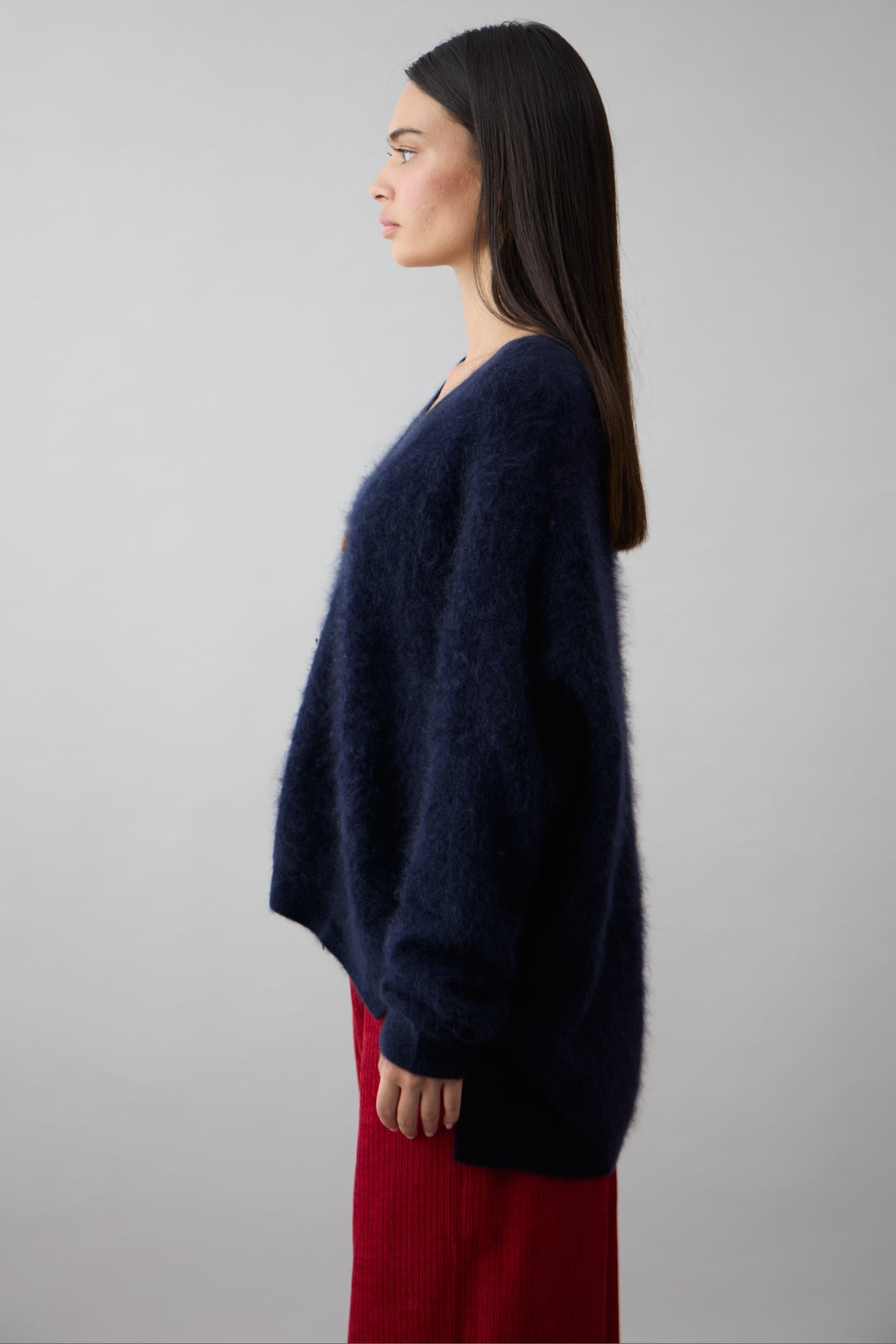 A woman with straight, dark hair stands in profile against a plain light background, wearing the navy Mae Cardi sweater by Mia Fratino Cashmere and red pants.