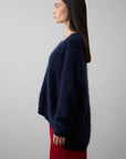 A woman with straight, dark hair stands in profile against a plain light background, wearing the navy Mae Cardi sweater by Mia Fratino Cashmere and red pants.