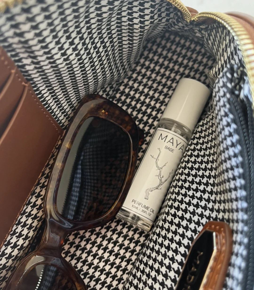 A close-up of a brown bag with black and white houndstooth lining holds tortoiseshell sunglasses and a bottle of Maya Fragrances' MAYA BASE 10 vegan, cruelty-free fragrance oil.