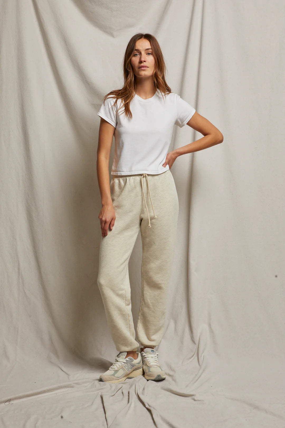 A person stands in front of a neutral draped background, wearing a white T-shirt, Michelle Fleece Jogger by Perfectwhitetee in beige with a braided drawcord, and white sneakers. Their hair is long and loose, one hand on their hip, showcasing a relaxed posture.