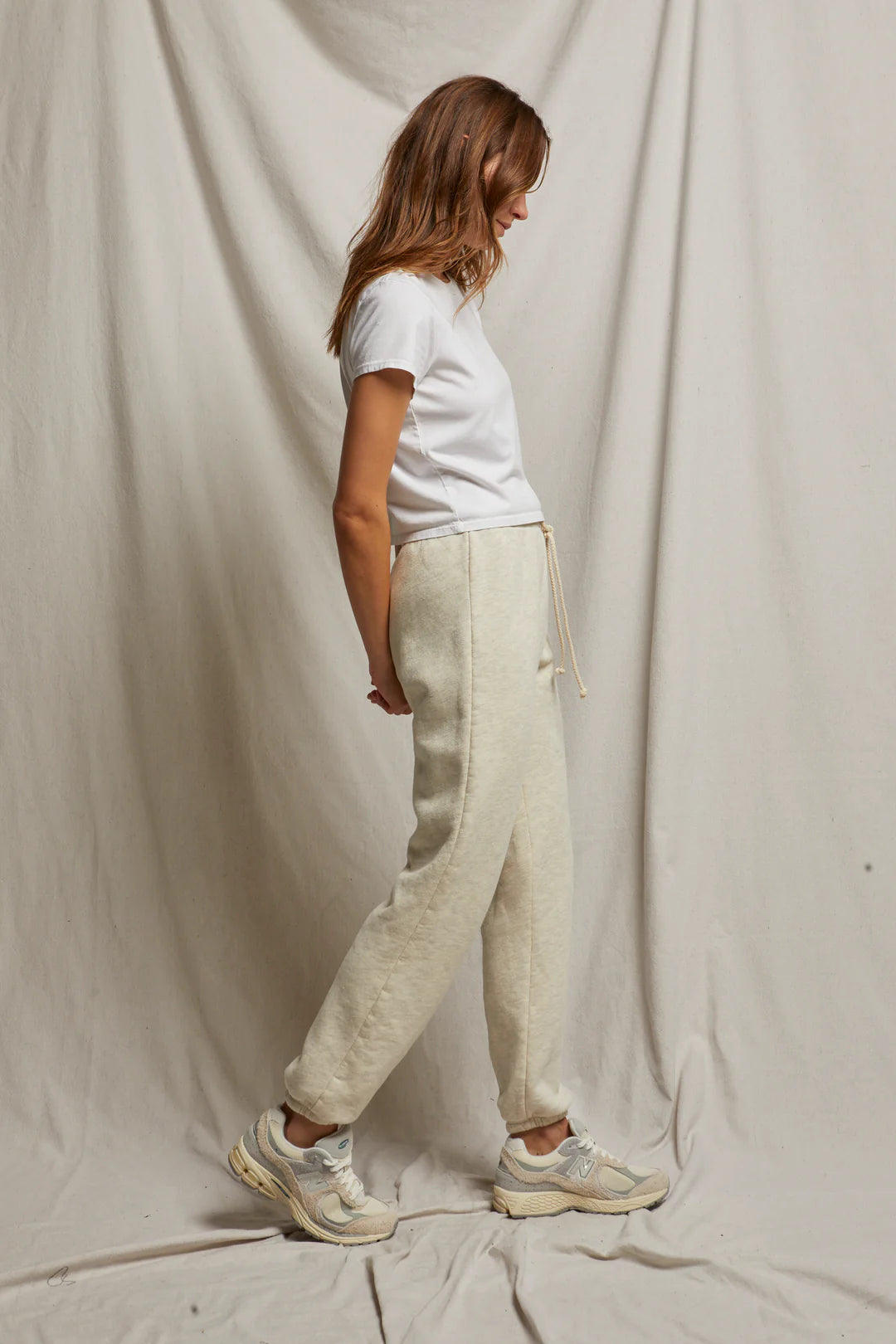 A person with long hair is standing sideways against a light fabric backdrop, highlighting the relaxed fit of the Michelle Fleece Jogger in beige. They pair it with a white t-shirt and white sneakers, enhancing the casual vibe and showcasing Perfectwhitetee's comforting sweatpant silhouette.