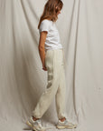 A person with long hair is standing sideways against a light fabric backdrop, highlighting the relaxed fit of the Michelle Fleece Jogger in beige. They pair it with a white t-shirt and white sneakers, enhancing the casual vibe and showcasing Perfectwhitetee's comforting sweatpant silhouette.