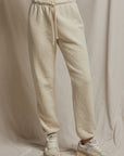 A person dressed in the Michelle Fleece Jogger from Perfectwhitetee with a braided draw cord and white sneakers stands against a beige fabric backdrop. The joggers showcase a classic silhouette, while the sneakers have light gray accents. The setting is simple and neutral, emphasizing subtle style.