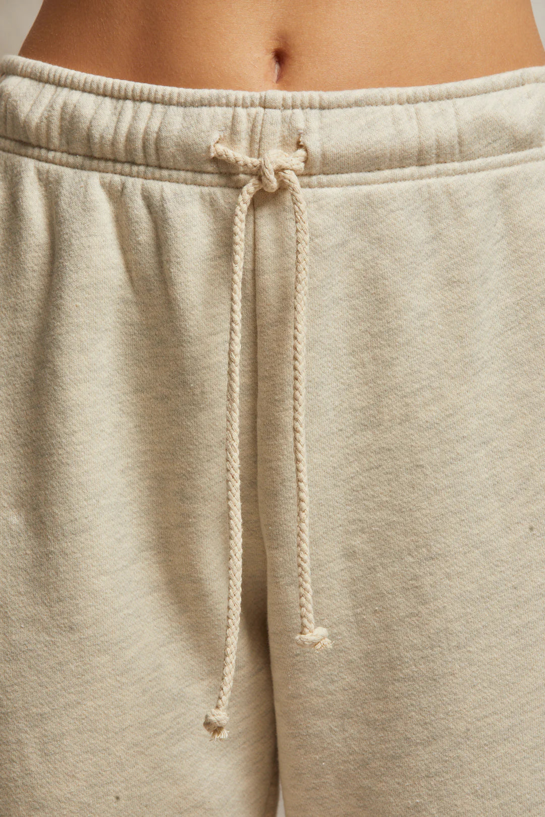 A close-up shows an individual wearing the Michelle Fleece Jogger by Perfectwhitetee, in a relaxed fit with beige fabric. The joggers feature a braided drawcord tied at the waist, and their cotton fleece material looks soft and comfortable with a slightly loose fit. The background is neutral and blurred.