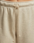 A close-up shows an individual wearing the Michelle Fleece Jogger by Perfectwhitetee, in a relaxed fit with beige fabric. The joggers feature a braided drawcord tied at the waist, and their cotton fleece material looks soft and comfortable with a slightly loose fit. The background is neutral and blurred.