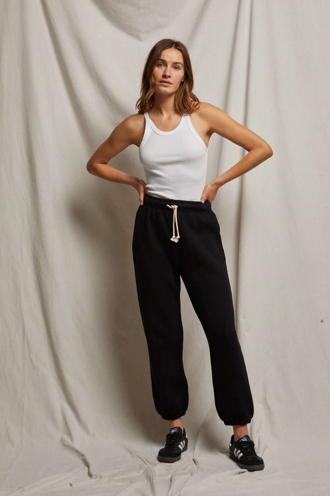 A person stands against a draped fabric background, wearing a white tank top and the Michelle Fleece Jogger from Perfectwhitetee, featuring a sweatpant silhouette with a relaxed fit, paired with black sneakers. With hands on hips, they exude effortless ease.