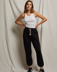 A person stands against a draped fabric background, wearing a white tank top and the Michelle Fleece Jogger from Perfectwhitetee, featuring a sweatpant silhouette with a relaxed fit, paired with black sneakers. With hands on hips, they exude effortless ease.