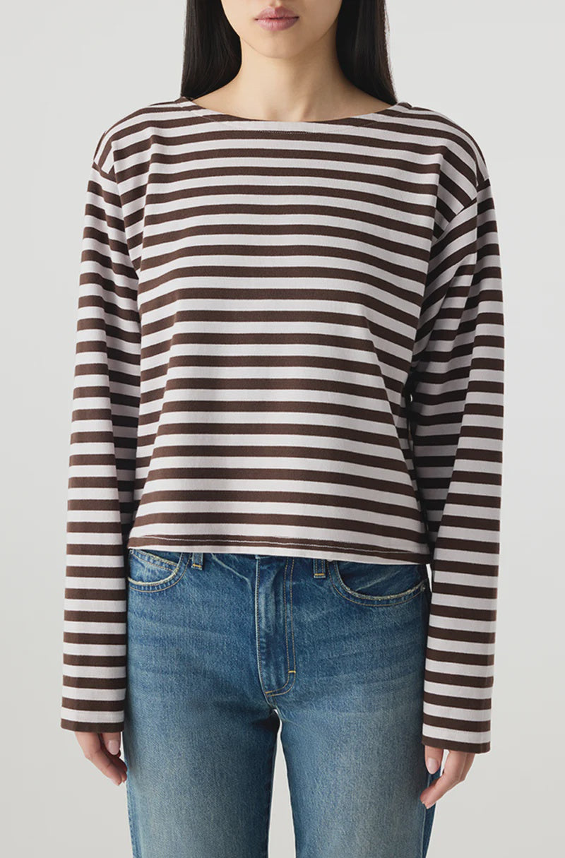 A woman wears the AMO Denim Hepburn Top, a relaxed fit, heavyweight cotton long sleeve tee with brown and white stripes, paired with blue jeans. She stands against a plain light background with her face partially visible and arms by her sides.