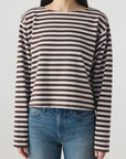 A woman wears the AMO Denim Hepburn Top, a relaxed fit, heavyweight cotton long sleeve tee with brown and white stripes, paired with blue jeans. She stands against a plain light background with her face partially visible and arms by her sides.