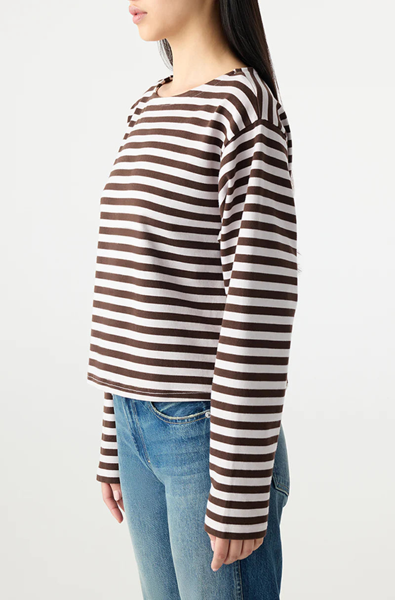 A woman stands in profile wearing the Hepburn Top by AMO Denim, a relaxed fit heavyweight cotton long sleeve tee with brown and white stripes, paired with light blue jeans against a plain white background.
