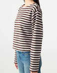 A woman stands in profile wearing the Hepburn Top by AMO Denim, a relaxed fit heavyweight cotton long sleeve tee with brown and white stripes, paired with light blue jeans against a plain white background.