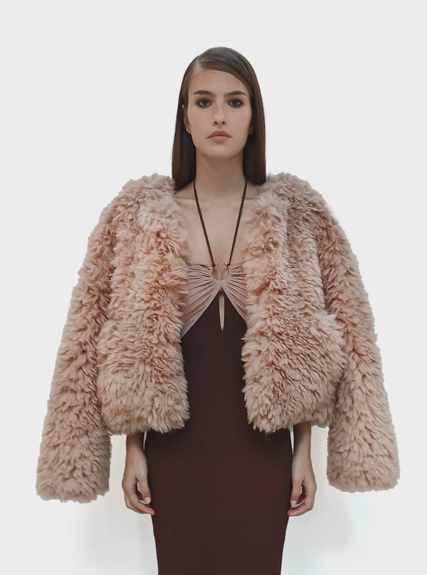 A woman with straight brown hair wears the Le Superbe Madison Chubby jacket—an oversized, light pink faux shearling style—over a fitted, sleeveless brown dress, standing against a plain white background.