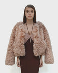 A woman with straight brown hair wears the Le Superbe Madison Chubby jacket—an oversized, light pink faux shearling style—over a fitted, sleeveless brown dress, standing against a plain white background.