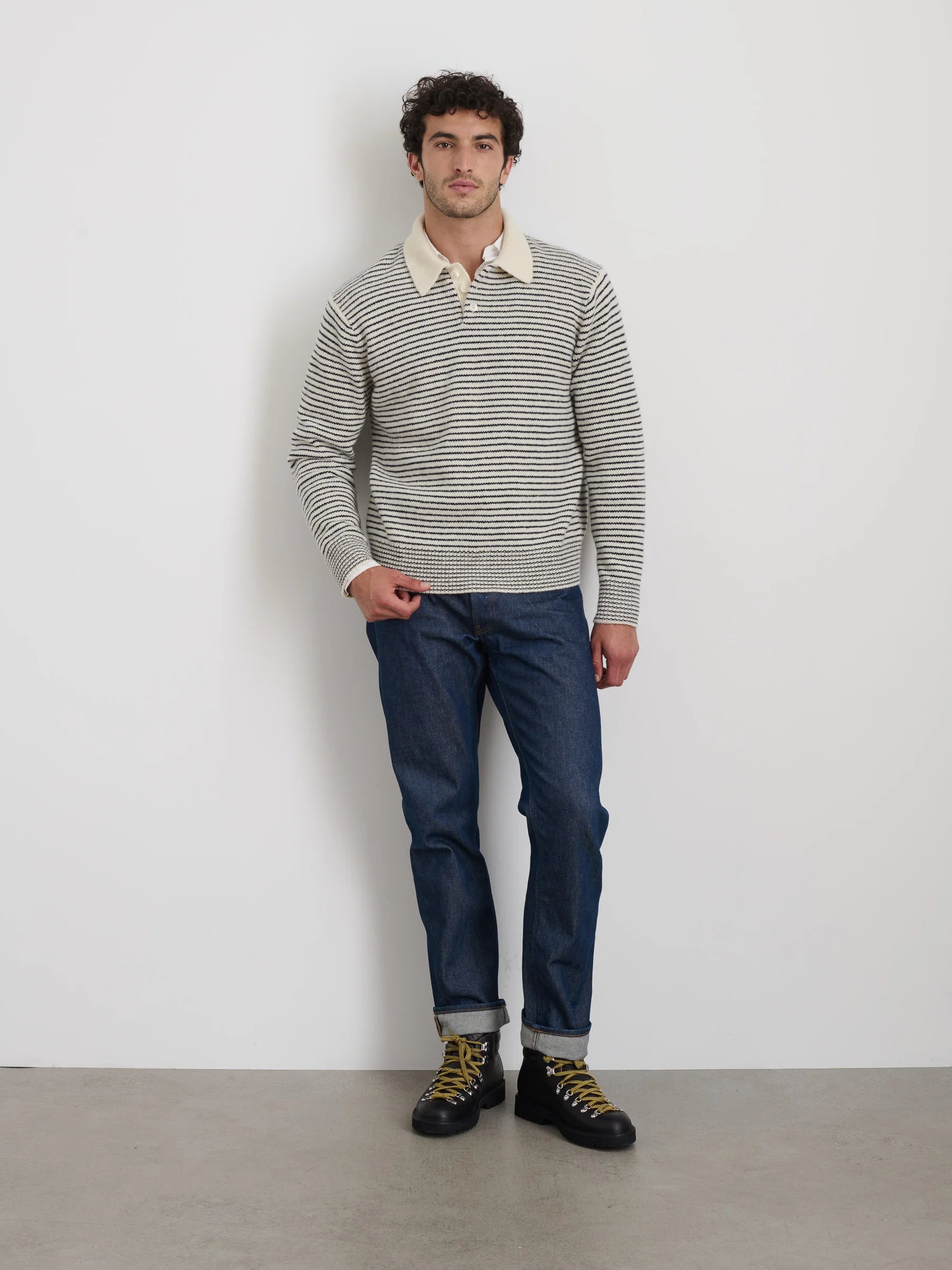 A man is standing against a white wall, wearing the Highland Collared Sweater by Alex Mill, blue jeans, and black boots with yellow laces.