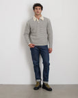 A man is standing against a white wall, wearing the Highland Collared Sweater by Alex Mill, blue jeans, and black boots with yellow laces.