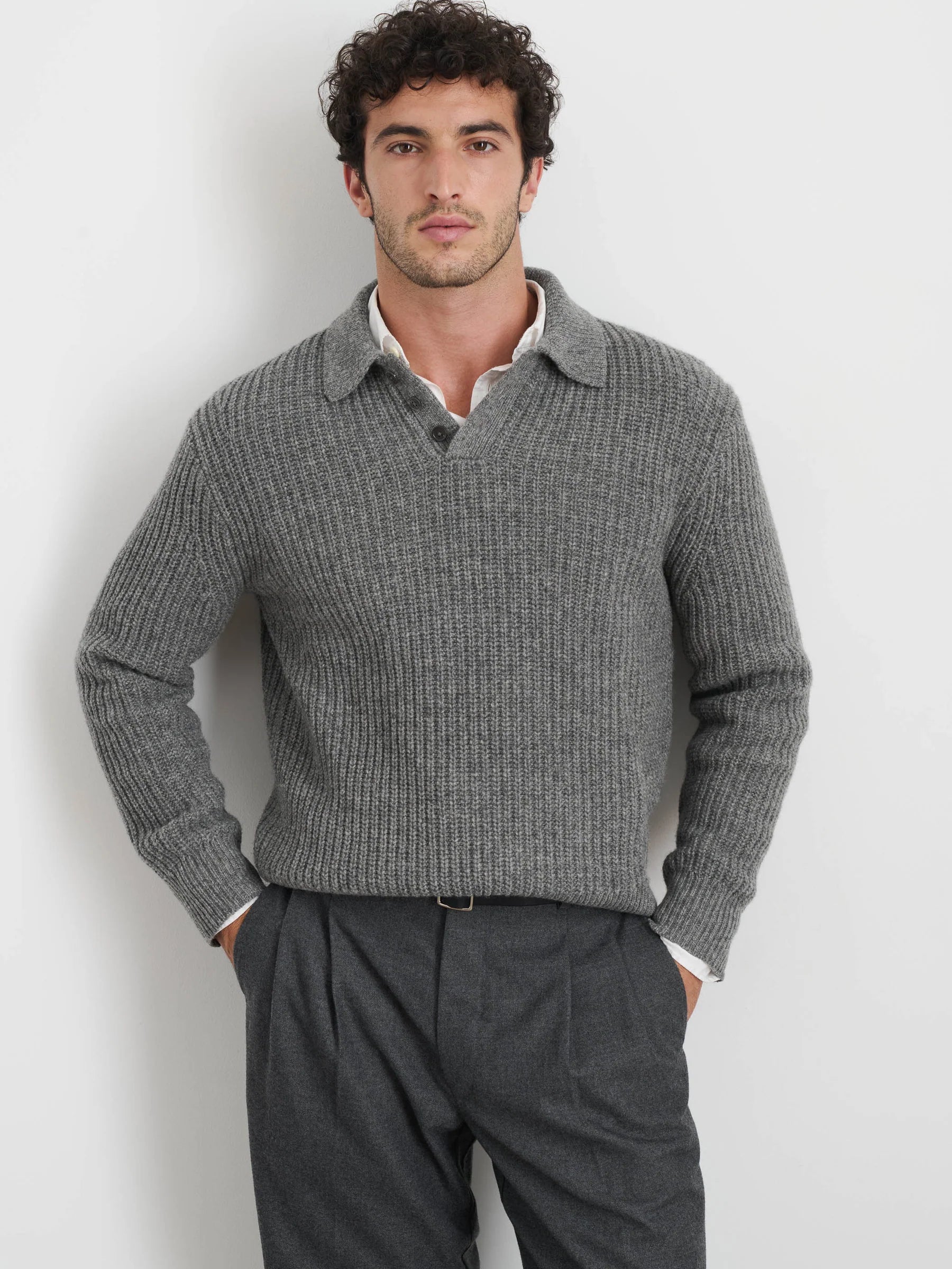 A man with curly hair stands against a plain background, wearing the Highland Collared Sweater by Alex Mill over a white collared shirt and dark gray pants. His hands are in his pockets as he looks directly at the camera.