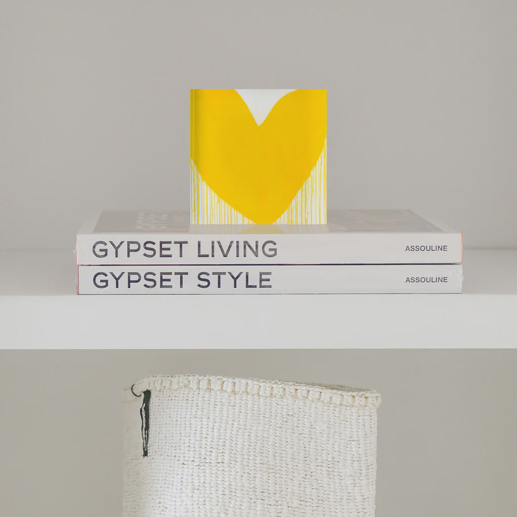 A small decorative item with a large yellow heart, aptly named "Mad Love Drippy Heart Block of Love" by Kerri Rosenthal, sits atop two stacked books titled "Gypset Living" and "Gypset Style" by Assouline. Below the shelf, a woven white basket is partially visible against the plain, light grey wall.