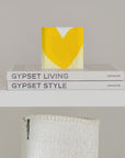 A small decorative item with a large yellow heart, aptly named "Mad Love Drippy Heart Block of Love" by Kerri Rosenthal, sits atop two stacked books titled "Gypset Living" and "Gypset Style" by Assouline. Below the shelf, a woven white basket is partially visible against the plain, light grey wall.