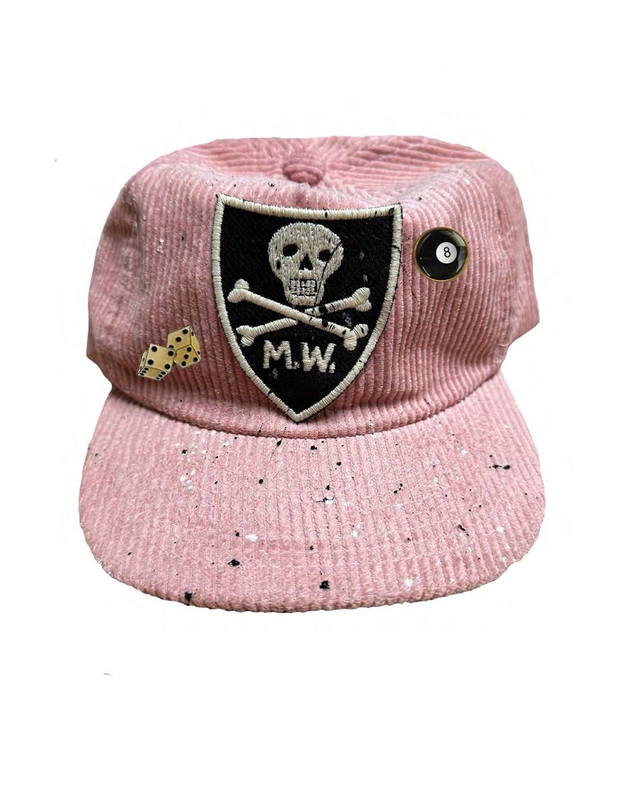 The MW Skull and Bones Hat by Made Worn is a pink corduroy hat with a black and white skull patch labeled "M.W.," dice and eight ball pins, plus black speckled paint details—perfect for your next field trip.