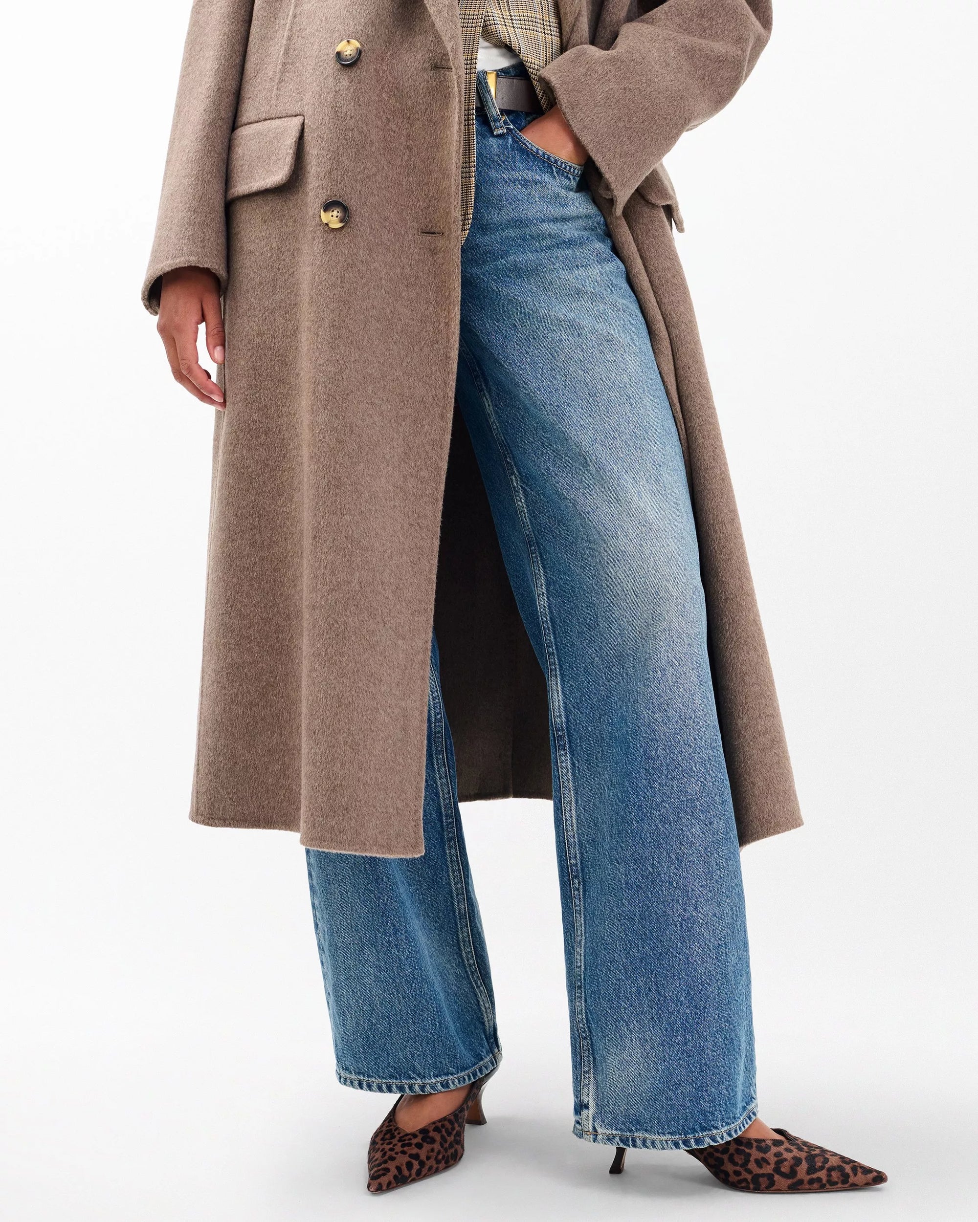 A person wears a long taupe coat over blue wide-leg jeans and a plaid shirt, with their left hand in their pocket and pointed leopard-print Rag & Bone madison slingback heels. The face is not visible.