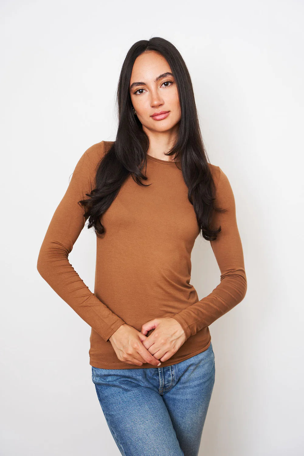 A woman with long dark hair wears a Majestic Filatures Soft Touch L/S Crewneck T-Shirt in brown and blue jeans, standing against a plain white background and looking at the camera with a relaxed expression.