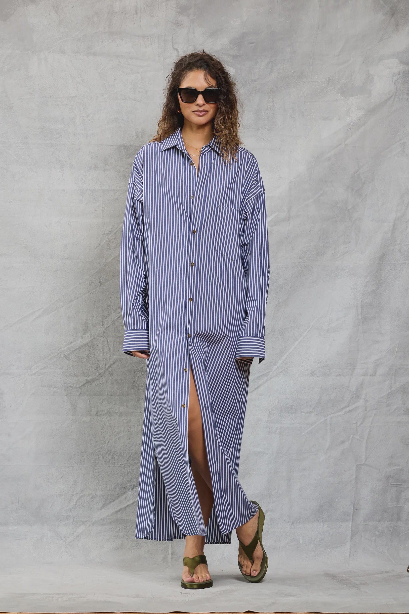 A woman in sunglasses and green sandals wears the Mamene Slim Phoebe Dress, a blue-and-white striped cotton shirtdress, standing against a light gray textured backdrop with one leg slightly forward.