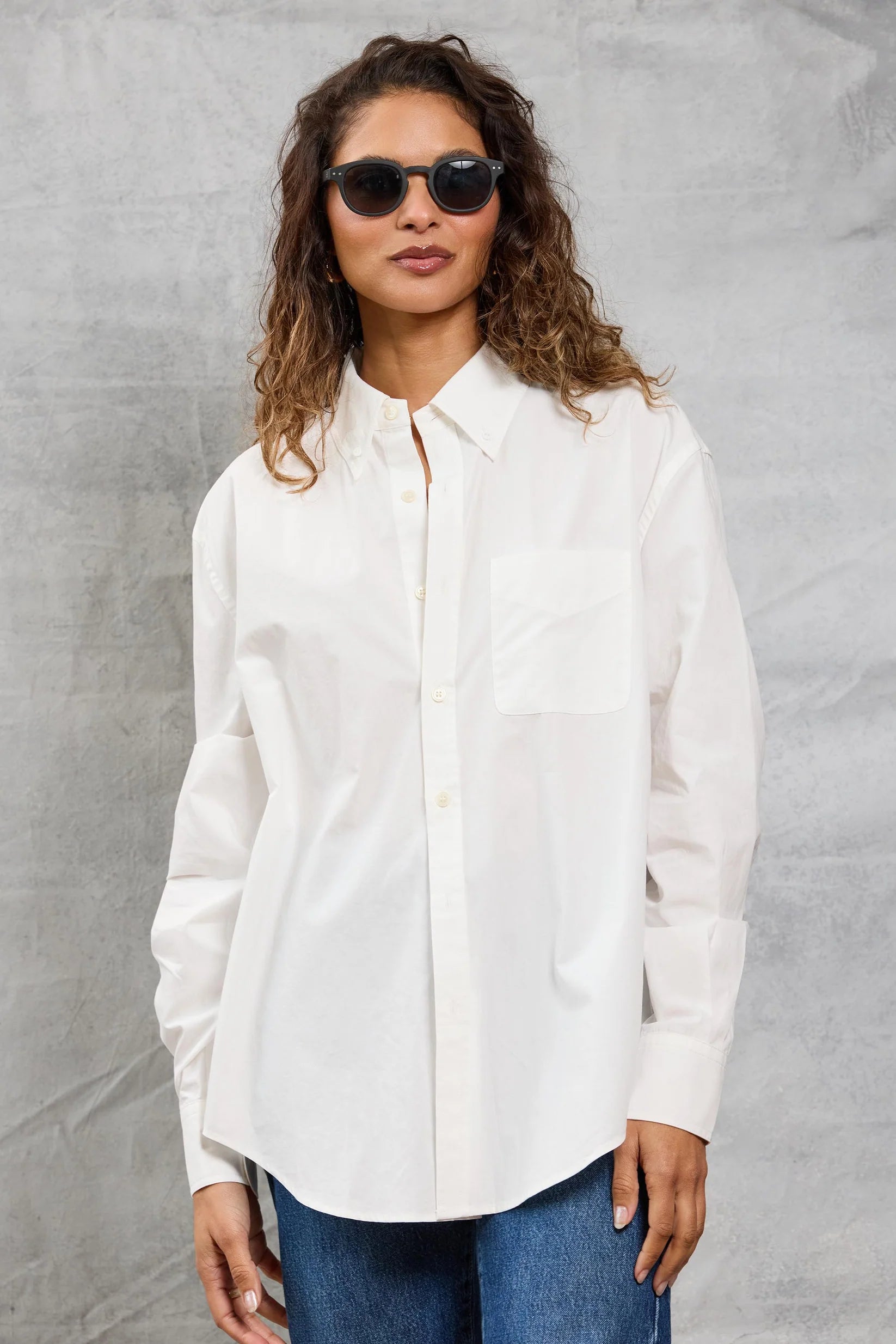 A woman with wavy brown hair and black sunglasses wears the Mamene Slim Repeat Shirt and blue jeans, standing against a light gray textured background.