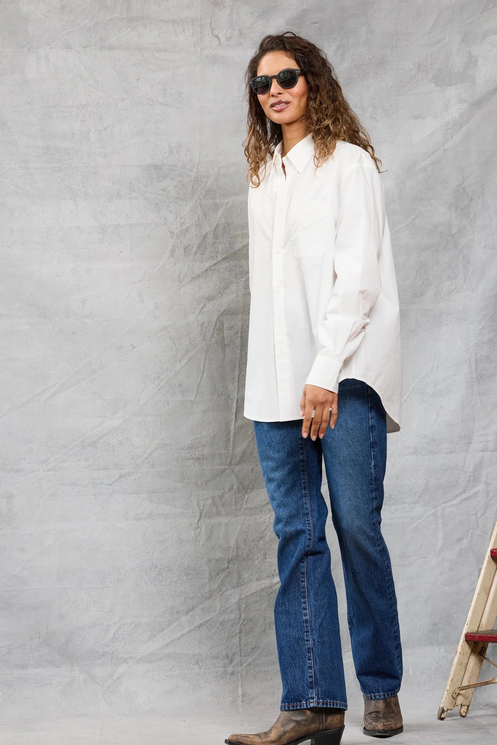 A person with long curly hair, sunglasses, blue jeans, and brown boots models the Mamene Slim Repeat Shirt against a light gray textured background near a wooden ladder.