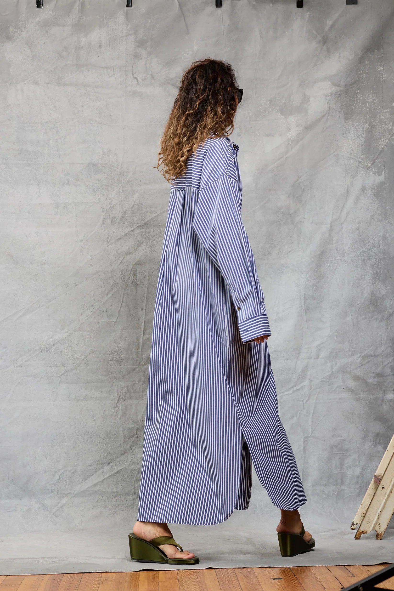 A woman with curly hair and sunglasses walks on a wooden floor against a light backdrop, wearing Mamene’s Slim Phoebe Dress—an ankle-length, navy and white striped cotton shirtdress—with green wedge sandals.