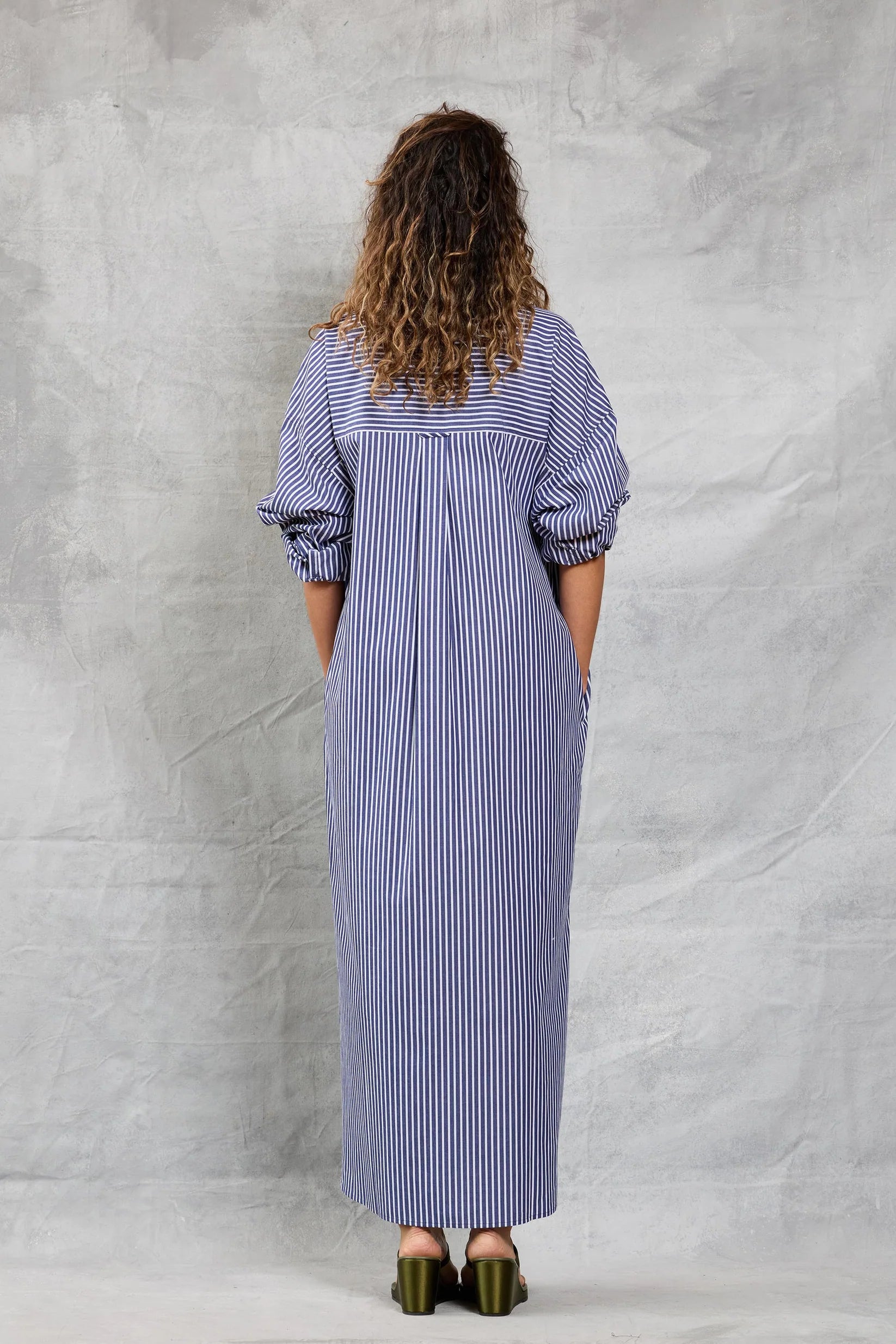 A person with curly hair faces away, wearing Mamene's Slim Phoebe Dress—an ankle-length, navy and white striped cotton shirtdress with rolled-up sleeves—and olive green heeled sandals against a textured light gray background.