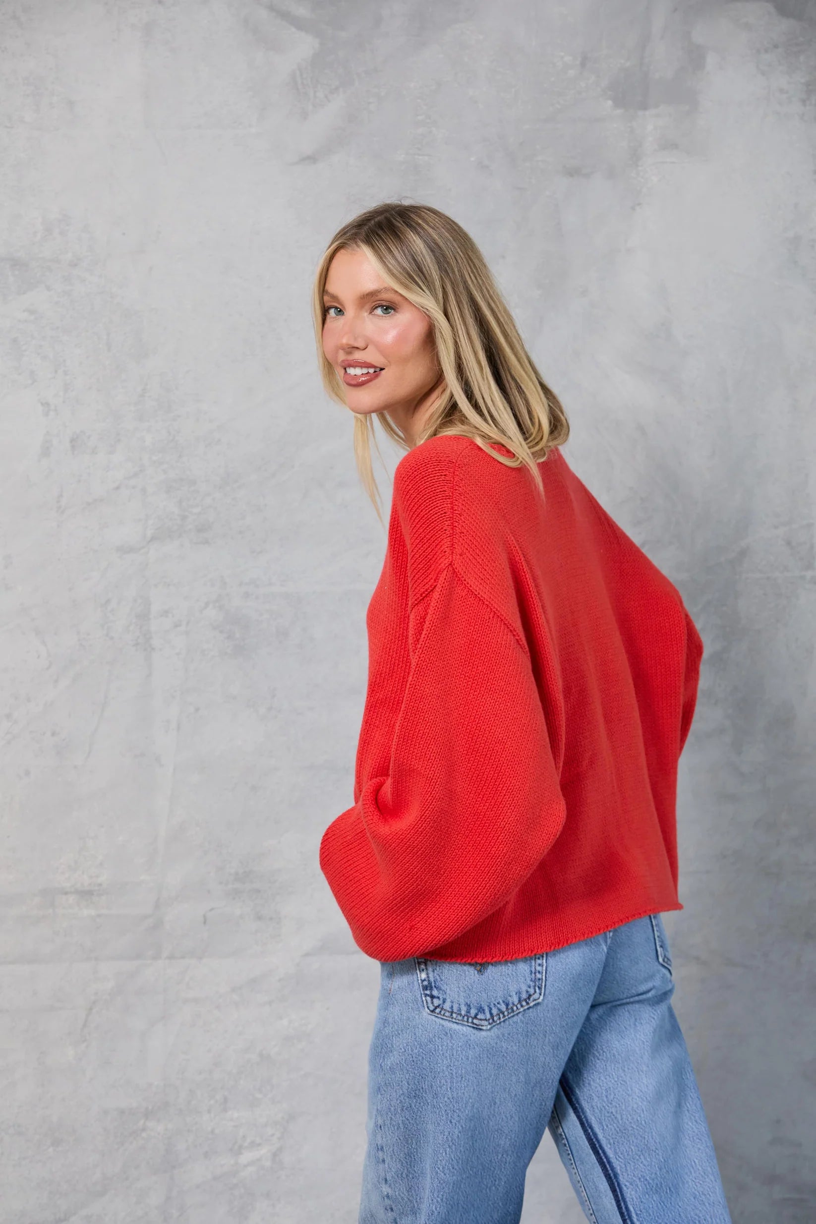 A woman with straight blonde hair stands sideways against a light gray textured backdrop, smiling over her shoulder while wearing the loose, oversized red Mamene Winona Sweater and blue jeans with her hands in her pockets.