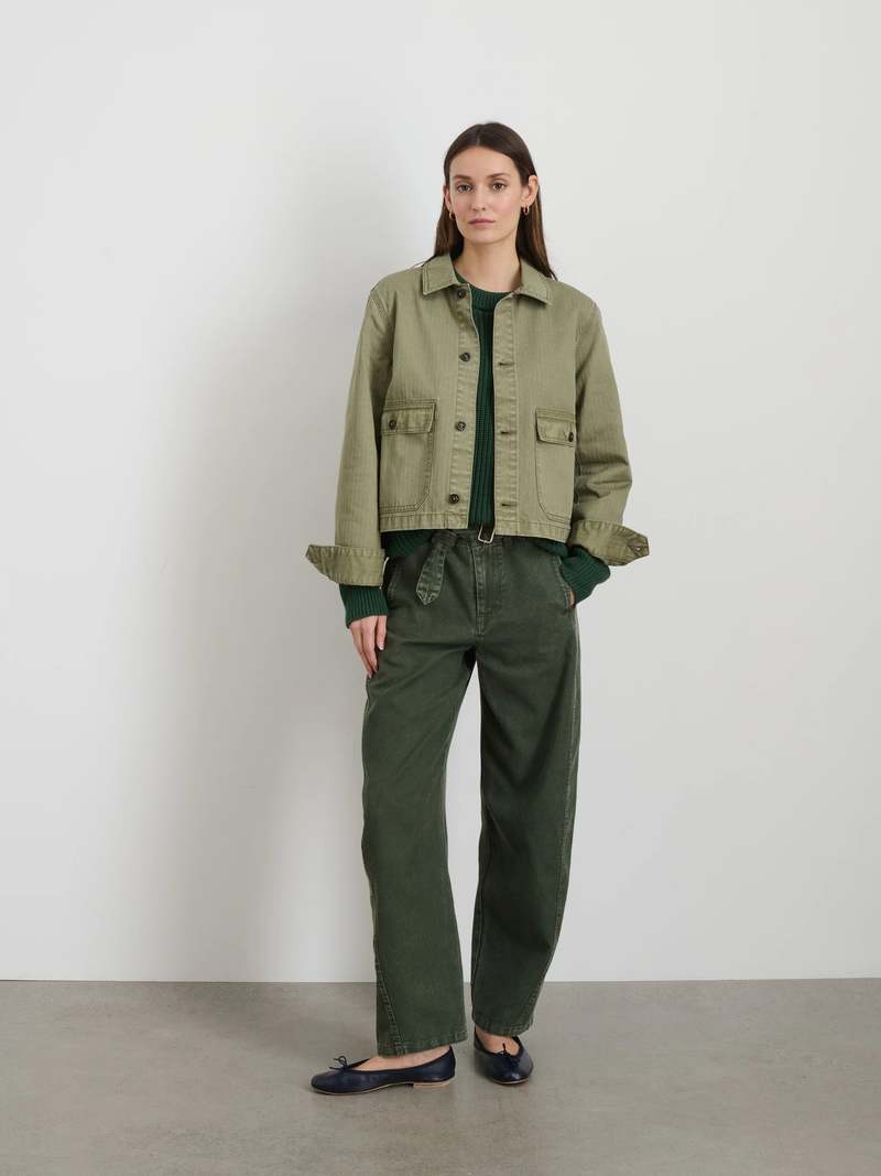 A woman with straight brown hair stands against a white wall, wearing the Alex Mill Marfa Work Jacket in garment-dyed olive green, paired with a green sweater, loose dark green pants, and black flats. Her hands are tucked into her pockets.