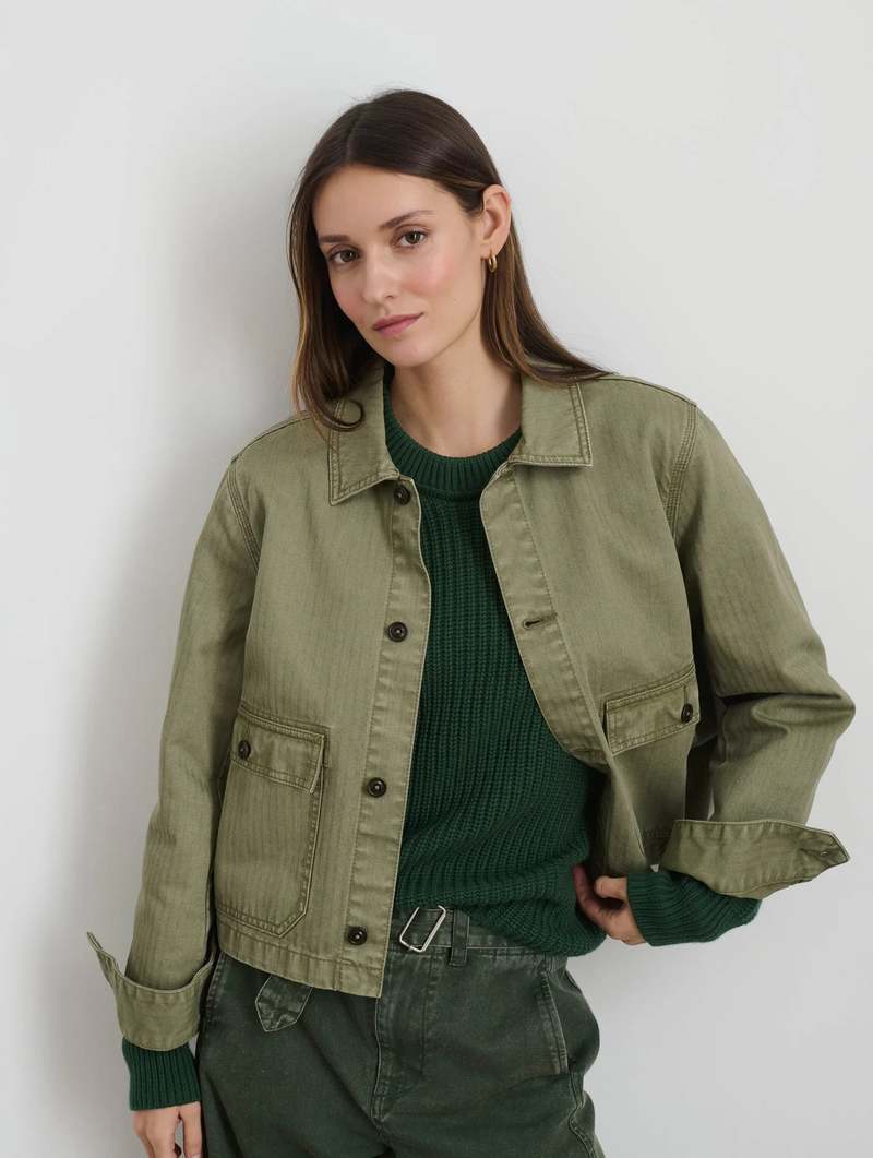 A woman with long brown hair wears the Marfa Work Jacket by Alex Mill in garment-dyed olive green over a dark green sweater and matching pants, standing against a white background with a relaxed expression.