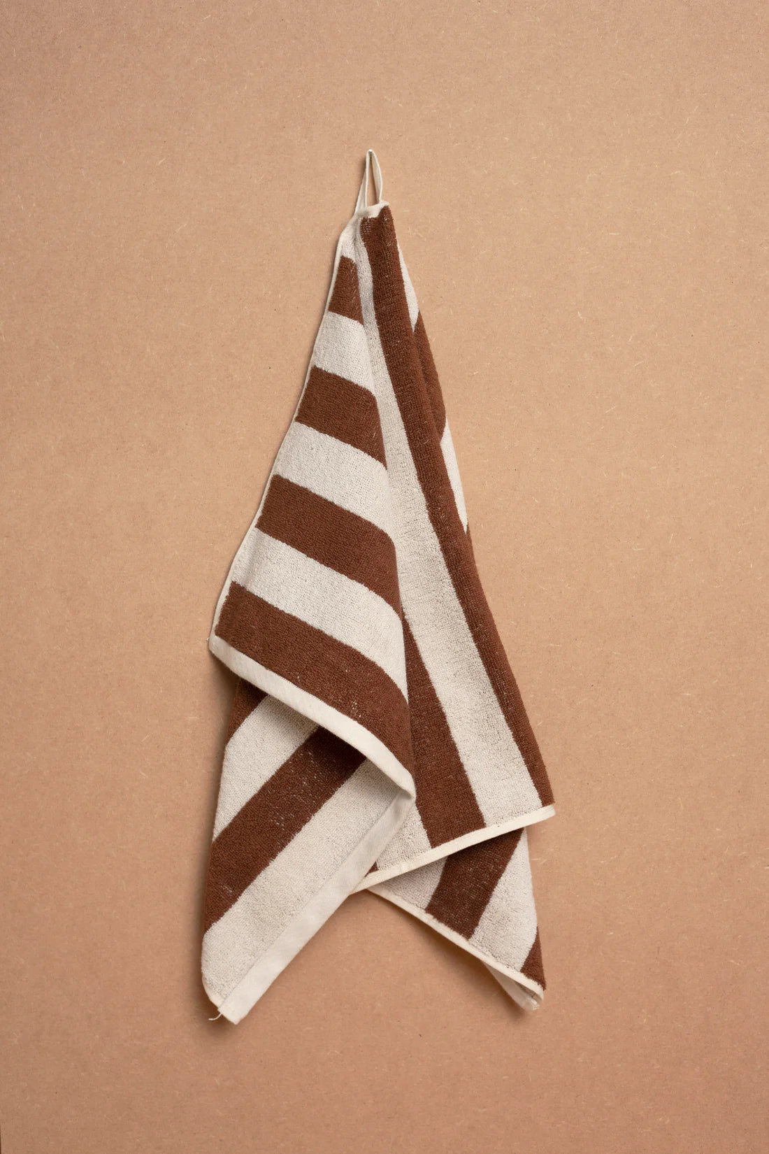The Maria Hand Towel by Autumn Sonata, featuring brown and white stripes, hangs on a hook against a tan wall. Crafted from soft organic cotton, it offers everyday comfort and style.