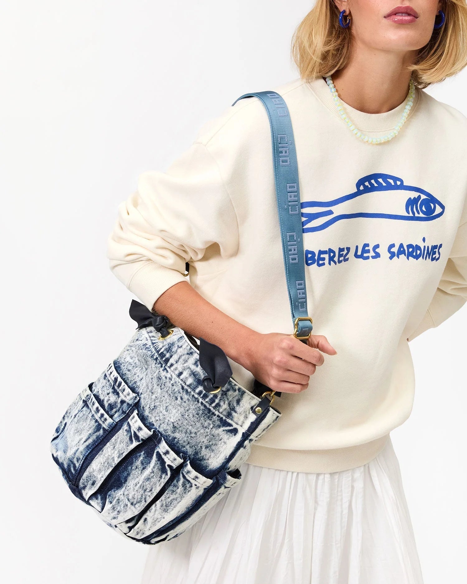 A woman wears a cream sweatshirt with a blue fish graphic and "LIBEREZ LES SARDINES" text, a white skirt, and carries the Clare Vivier Marine Bag—an acid wash denim purse shaped like jeans. She has short blonde hair and blue earrings.