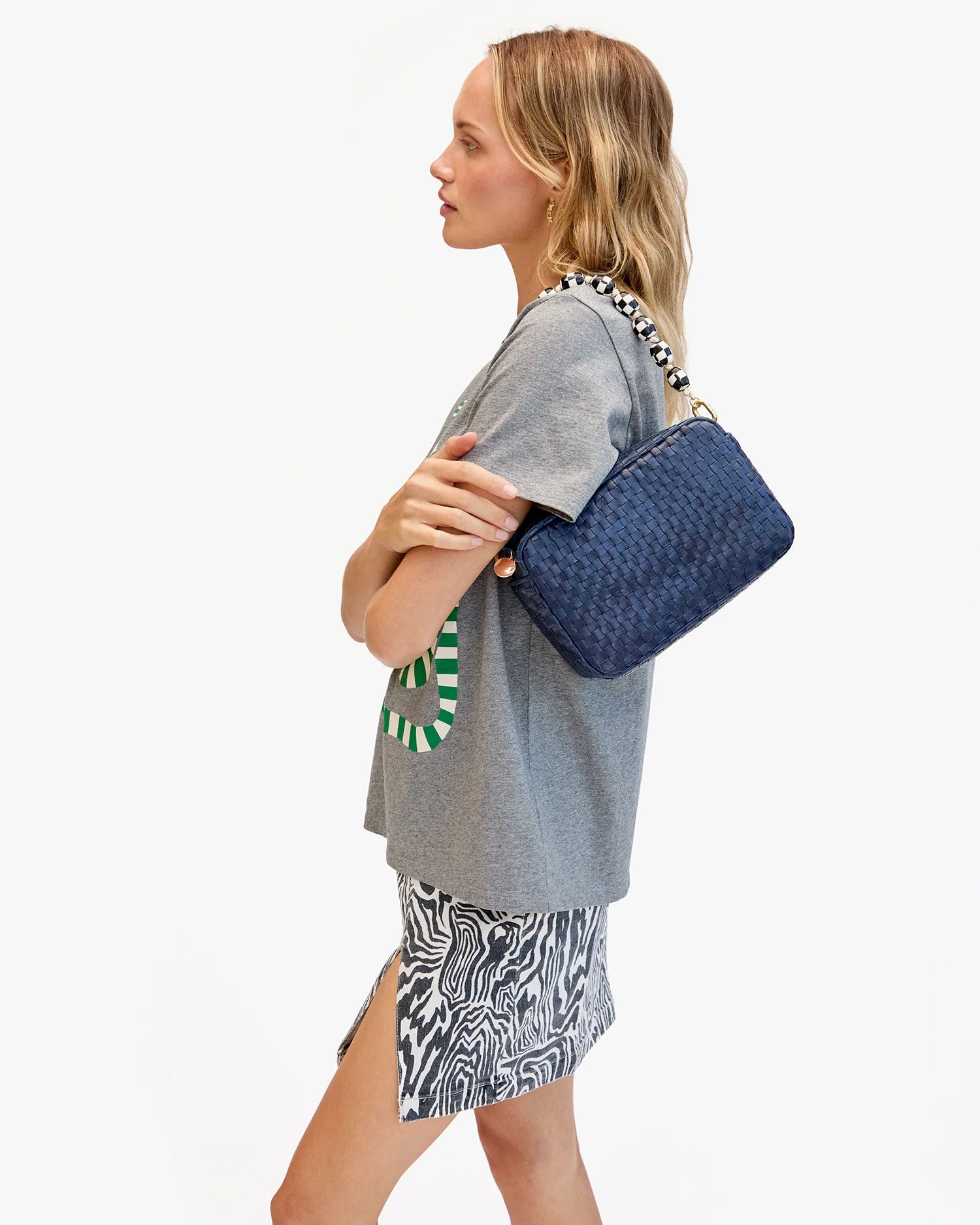 A woman in a gray graphic tee and black-and-white skirt stands in profile, holding the Clare Vivier Marisol navy blue purse with a crossbody strap. The background is plain white.
