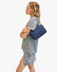 A woman in a gray graphic tee and black-and-white skirt stands in profile, holding the Clare Vivier Marisol navy blue purse with a crossbody strap. The background is plain white.