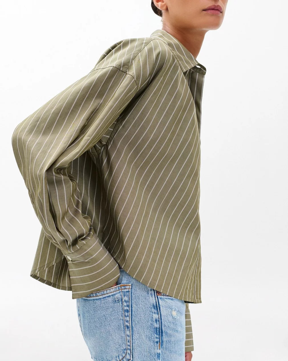 A person wears the Rag & Bone martha poplin striped shirt, a boxy fit olive-green top with white vertical stripes, paired with light blue high-waisted jeans against a plain white background.