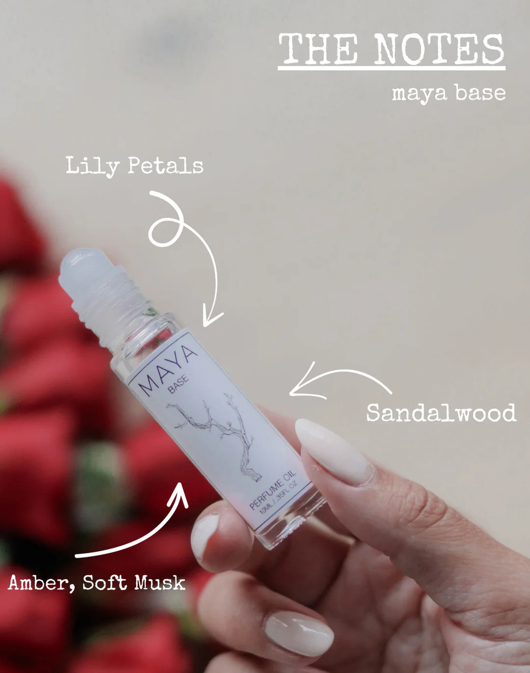 A hand holds a MAYA BASE 10 roll-on by Maya Fragrances. Arrows highlight lily petals, sandalwood, amber, and soft musk notes. Red flowers blur behind. Text reads “THE NOTES maya base.” Vegan and cruelty-free.