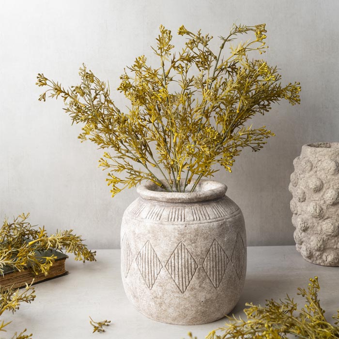 The Abigail Ahern Meadow Grass Flaxen is arranged in a textured ceramic vase with a diamond pattern, displayed on a light surface beside similar decor and foliage against a neutral background.
