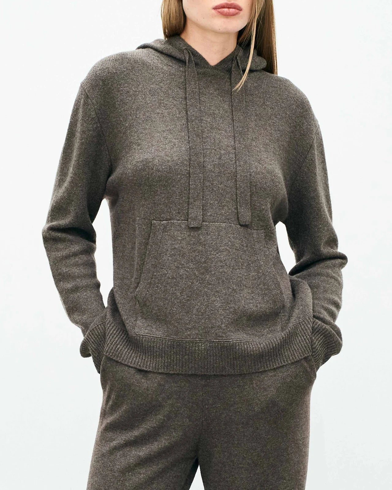 A woman stands with her hands in the pockets of a dark gray Jumper 1 2 3 4/CR2 Oversized Hoodie and matching pants against a light background. Her face is partially visible.