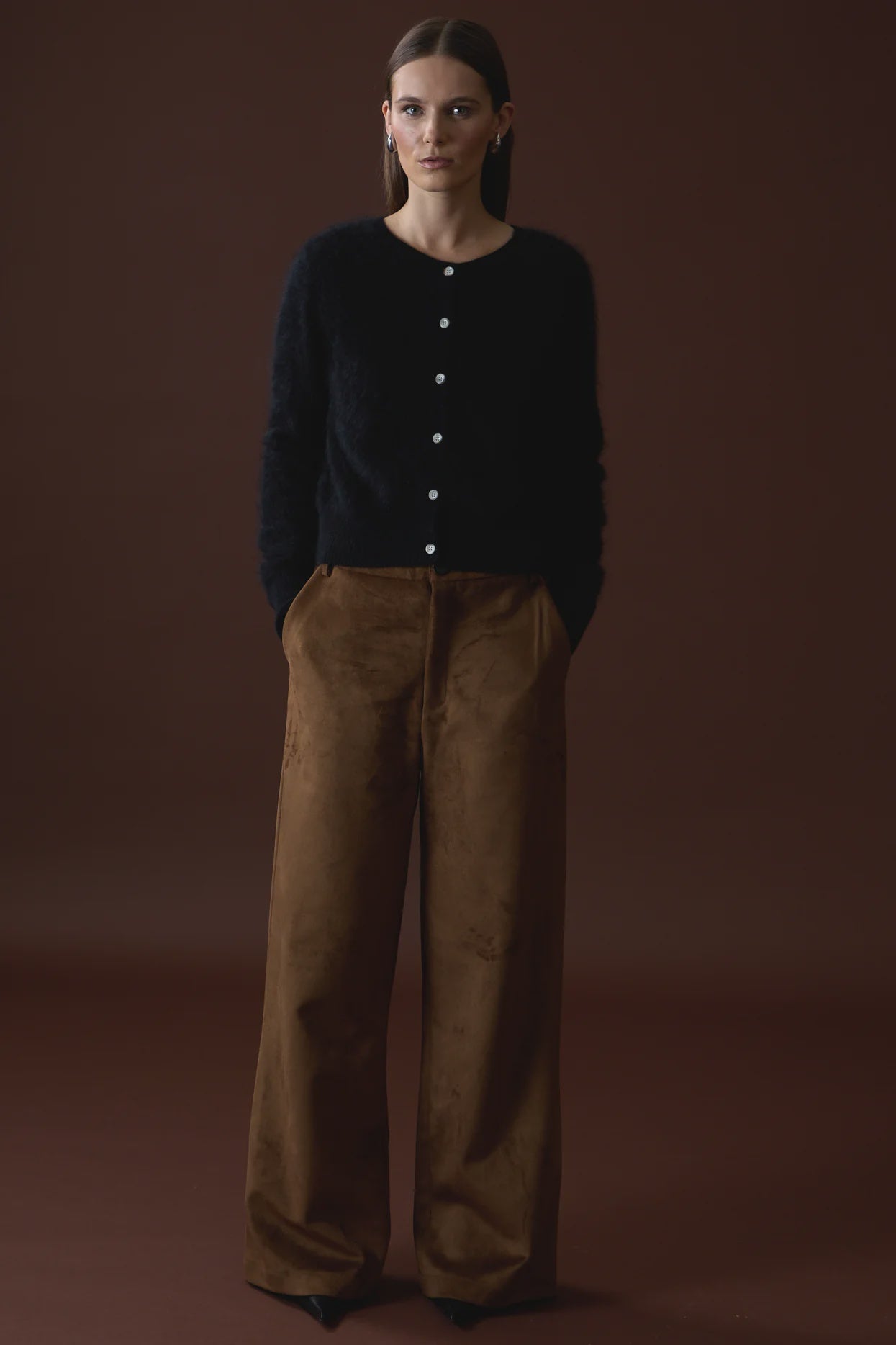 A woman with straight brown hair tucked behind her ears stands against a dark brown backdrop, hands in her pockets, wearing the Mia Fratino Cashmere Sienna Cardi and high-waisted, wide-leg brown pants.